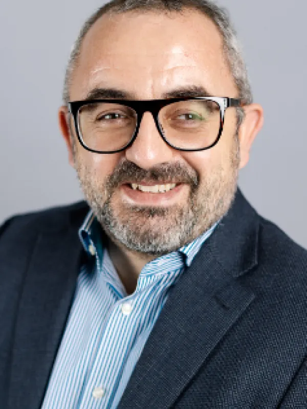 A front portrait of a man wearing a striped button down shirt and a blazer. He has big black glasses and a short beard.